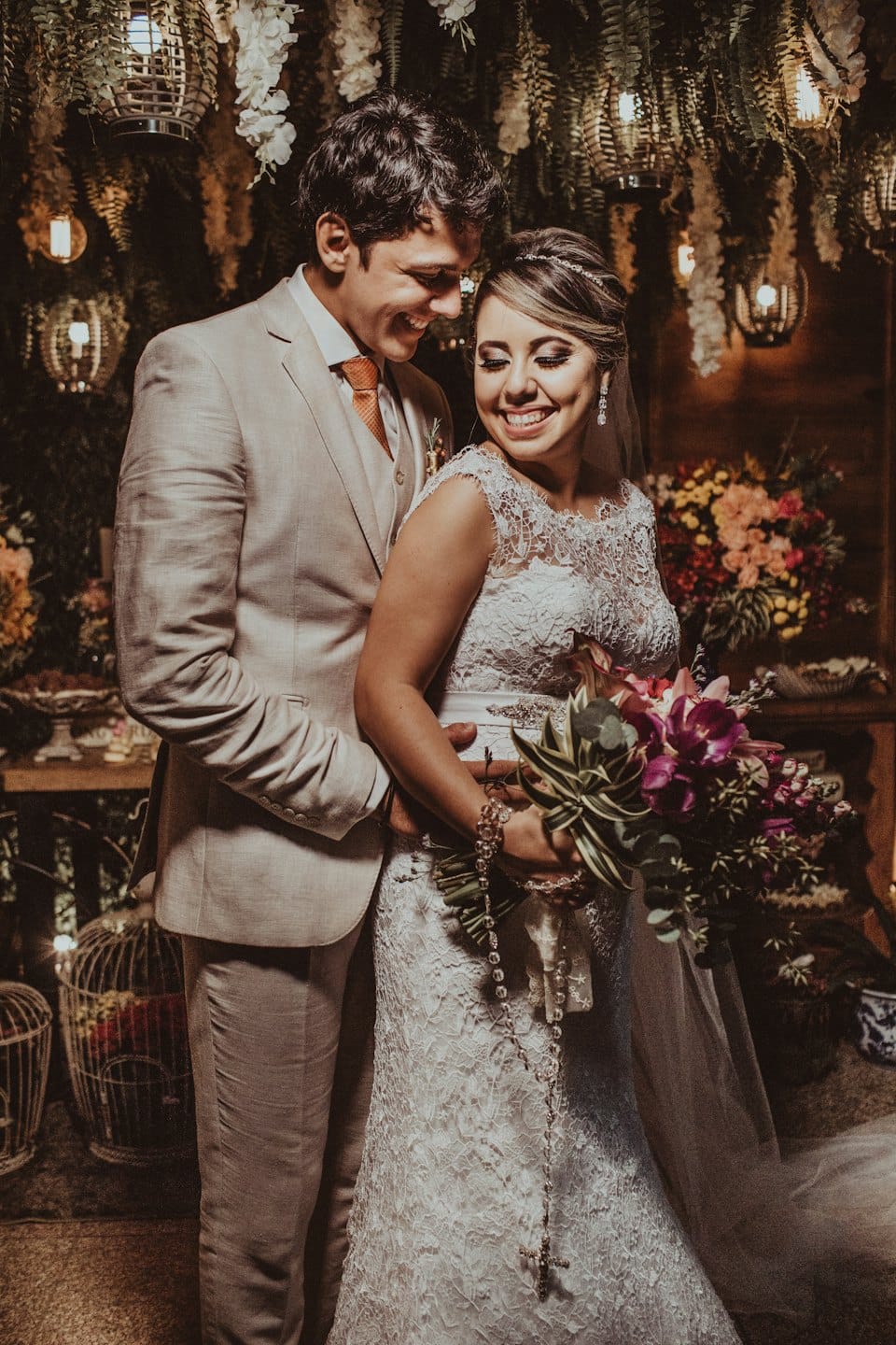Una pareja de recién casados se abraza rodeada de farolillos y flores colgantes.